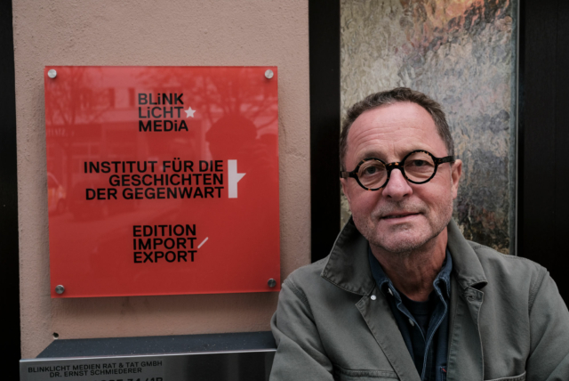 Ernst Schmiederer vor seinem Büro, rechts das Türschild mit Logos